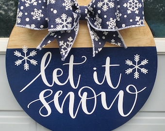 May include: A blue and brown round wooden door hanger with a white bow and snowflake pattern. The words "Let it snow" are written in white cursive lettering.