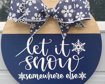 May include: A round wooden door hanger with a blue and white snowflake design. The top half of the hanger is natural wood and the bottom half is painted blue. A white ribbon with snowflakes is tied in a bow at the top. The text "Let it snow somewhere else" is written in white on the blue background.