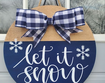 May include: A blue and brown round wooden door hanger with a white and black checkered bow and the text "Let it snow" in white cursive lettering with white snowflakes on either side of the text.