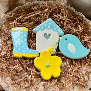 Puede incluir: Cuatro galletas de azúcar decoradas sobre un fondo de papel marrón. Las galletas son una bota de lluvia azul con lunares blancos, una casita de pájaros blanca con lunares azules, un pájaro azul con un ala blanca y una flor amarilla con un centro azul.