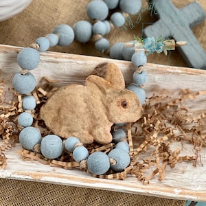 May include: A light blue beaded garland with a brown bunny figurine in a wooden tray with brown shredded paper. The garland is made of wooden beads and has a rustic look. The bunny is a vintage style toy.