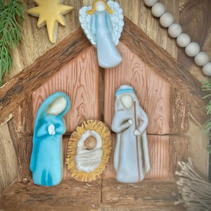 May include: A wooden nativity scene with a blue and white angel, a blue robed figure, a white robed figure, and a baby Jesus in a manger.