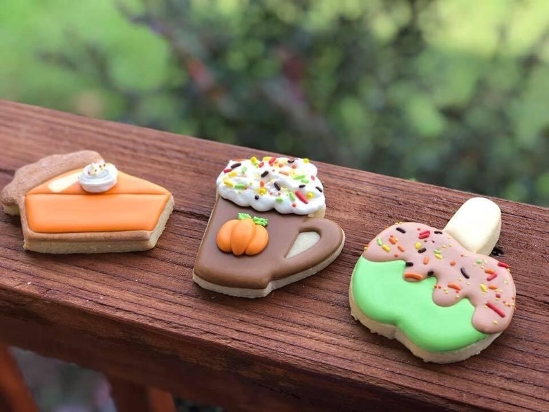 May include: Three decorated sugar cookies, a pumpkin pie, a pumpkin spice latte, and a caramel apple. The cookies are on a wooden surface.