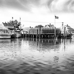 May include: Black and white photograph of a harbor scene. Fishing boats are docked at a wooden pier, with buildings and an American flag in the background. The water reflects the sky and structures.