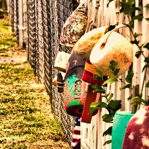 May include: A collection of weathered buoys in various colors, including red, green, and white, hang on a white picket fence. The buoys are attached with rope and are next to a chain-link fence. An American flag is visible.