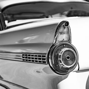 May include: Black and white image of a classic car's rear section. The focus is on the tail light assembly, featuring a circular design with a central lens and chrome accents. The car's bodywork is smooth and reflective.