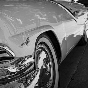 May include: Black and white image of a classic car's front quarter. The car has a chrome grill, headlight, and a white-walled tire. The body is a light color, and the car appears to be parked on a street.