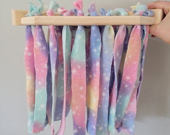 Puede incluir: Un juguete de madera con tiras de tela de colores arcoíris atadas a él. La tela tiene un patrón de estrellas blancas. El juguete está colgado de una pared.