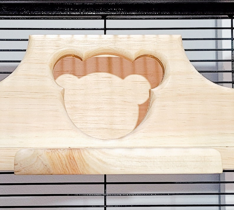 Puede incluir: Un estante de madera con un recorte en forma de cabeza de oso. El estante est&aacute; dise&ntilde;ado para encajar dentro de una jaula.
