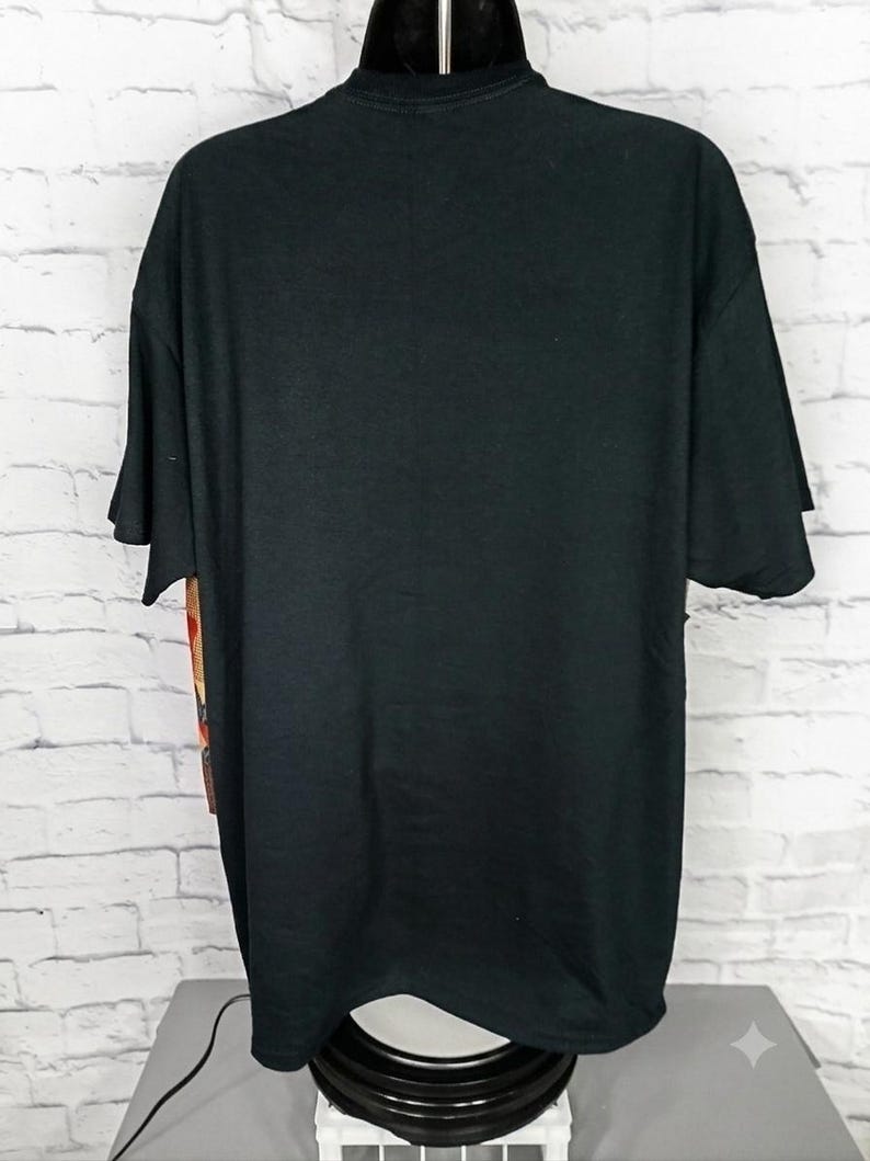 Może przedstawiać: Gładki czarny t-shirt z kr&oacute;tkim rękawem na manekinie. Odzież ma gładki tył z okrągłym dekoltem i standardowym obszyciem, z widocznym fragmentem graficznego nadruku na bocznym panelu koszulki.