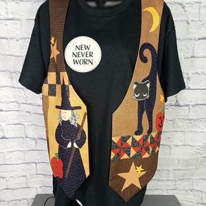 Może przedstawiać: Czarny t-shirt noszony pod bezrękawnikiem z motywem Halloween. Kamizelka przedstawia czarownicę, czarnego kota, p&oacute;łksiężyc, gwiazdy i dynię. Okrągła naklejka na t-shircie zawiera napis NEW NEVER WORN czarnymi literami.