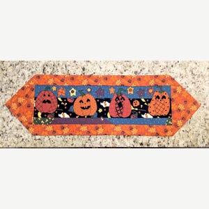 May include: A rectangular fabric table runner featuring a central horizontal panel with four orange jack-o'-lanterns on a dark blue background with stars and small pumpkins. The border is orange with a repeating pattern of small brown autumn leaves.