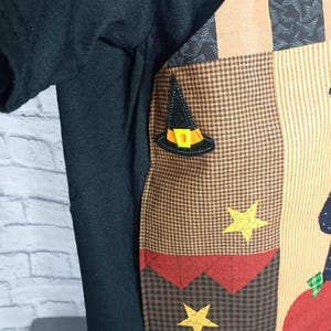 Może przedstawiać: Czarny t-shirt z naszytym panelem z tkaniny w stylu patchwork z przodu. Panel posiada brązowo-beżowy wz&oacute;r w kratę, aplikację czarnego kapelusza czarownicy z ż&oacute;łtą klamrą oraz dwie ż&oacute;łte gwiazdki. Widoczny jest r&oacute;wnież kształt czerwonego jabłka.