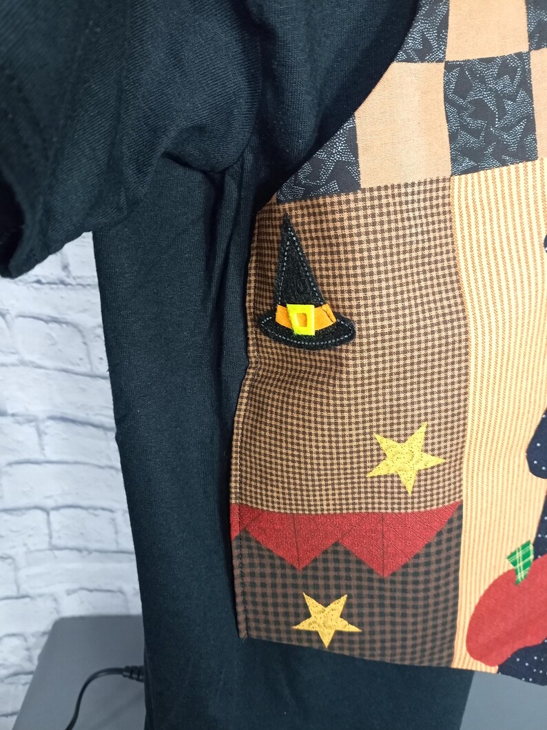 Może przedstawiać: Czarny t-shirt z naszytym panelem z tkaniny w stylu patchwork z przodu. Panel posiada brązowo-beżowy wz&oacute;r w kratę, aplikację czarnego kapelusza czarownicy z ż&oacute;łtą klamrą oraz dwie ż&oacute;łte gwiazdki. Widoczny jest r&oacute;wnież kształt czerwonego jabłka.