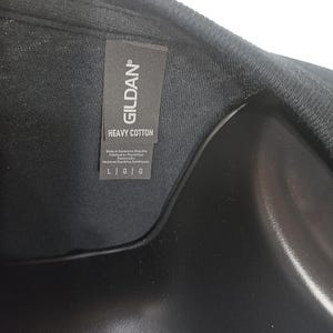Może przedstawiać: Czarny t-shirt z szarą metką materiałową przy kołnierzu, na kt&oacute;rej nadrukowano białą nazwę marki GILDAN oraz napis HEAVY COTTON. Metka zawiera r&oacute;wnież informacje o produkcji oraz rozmiar L G G wydrukowany wyraźną i czytelną czcionką.