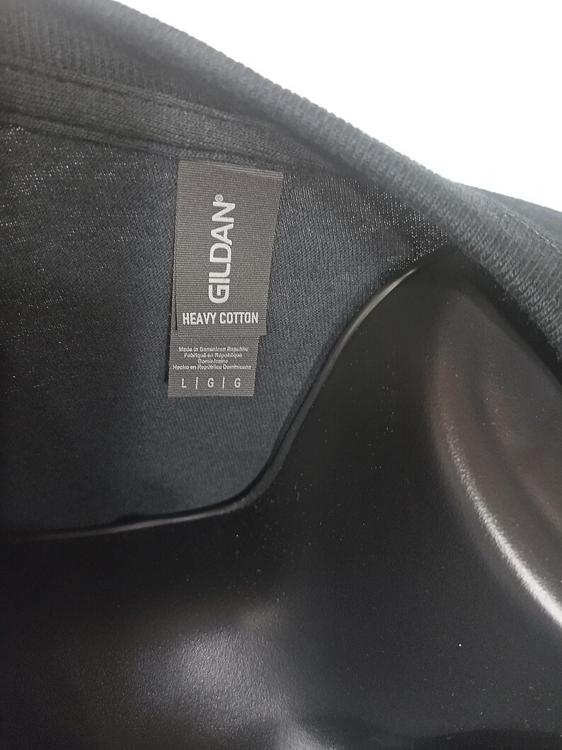 Może przedstawiać: Czarny t-shirt z szarą metką materiałową przy kołnierzu, na kt&oacute;rej nadrukowano białą nazwę marki GILDAN oraz napis HEAVY COTTON. Metka zawiera r&oacute;wnież informacje o produkcji oraz rozmiar L G G wydrukowany wyraźną i czytelną czcionką.