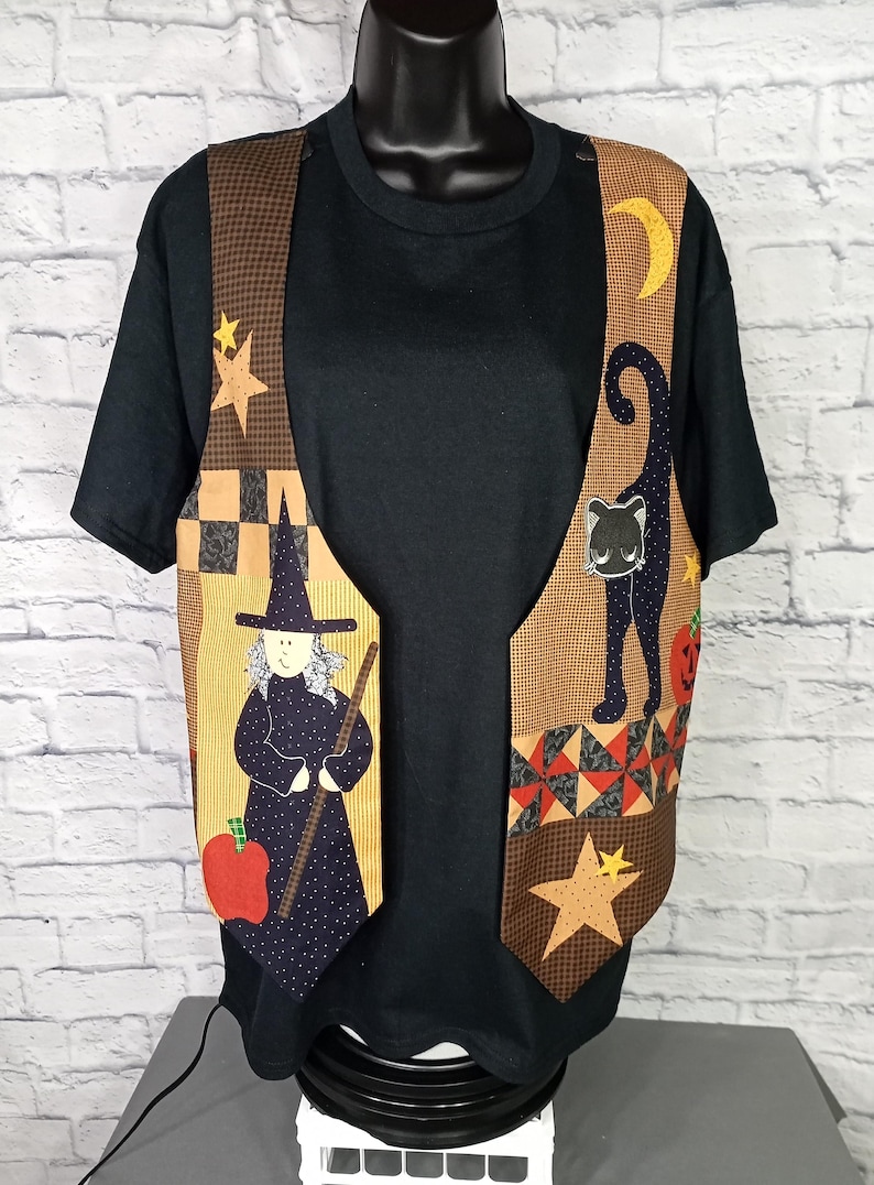 Może przedstawiać: Czarny t-shirt z kr&oacute;tkim rękawem noszony pod bezrękawnikiem z patchworkowym wzorem. Kamizelka przedstawia czarownicę z miotłą, czarnego kota, ż&oacute;łty p&oacute;łksiężyc, gwiazdy, czerwone jabłko i dynię na brązowym materiale.