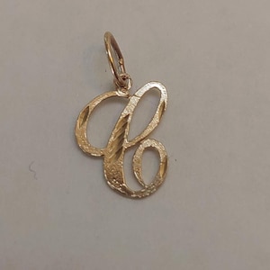 Puede incluir: Un colgante dorado con la forma de la letra "C" en escritura cursiva. El colgante tiene una superficie texturizada y un lazo en la parte superior para sujetarlo a una cadena. Probablemente de oro, una joya.