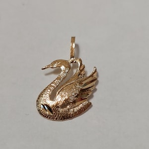 May include: A gold swan pendant with detailed craftsmanship. The pendant depicts a swan with a curved neck and textured wings. The swan's body has a textured surface. A small loop is at the top for attaching a chain.