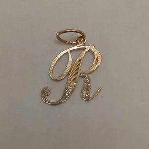 以下が含まれることがあります： 金色の「R」の文字のペンダント。ペンダントは、文字の本体にテクスチャ加工が施され、輪郭に滑らかで研磨された仕上げが施された筆記体のデザインです。チェーン用の小さなループが上部に取り付けられています。