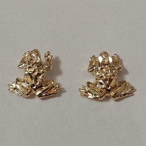 May include: A pair of gold frog earrings. The frogs are sitting on their hind legs with their front legs raised. The earrings are detailed with textured surfaces.