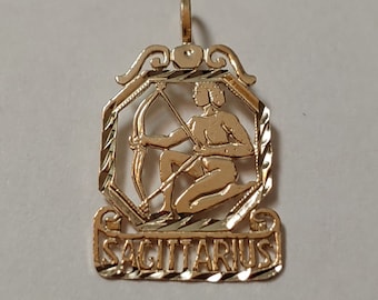 May include: Gold-toned pendant featuring a detailed depiction of the Sagittarius zodiac sign, with a figure holding a bow and arrow, and the word "SAGITTARIUS" inscribed below.