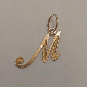 May include: A gold-colored pendant in the shape of the letter "M". The pendant has a textured surface and a loop at the top for a chain. The design is a cursive script style.
