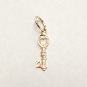 May include: A gold-colored key charm with a heart-shaped keyhole. The charm is designed to be used as a pendant or a charm for a bracelet.