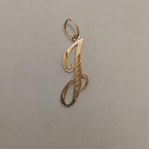 Op de afbeelding: Een goudkleurige hanger in de vorm van de letter "J". De hanger heeft een textuurontwerp en een kleine lus aan de bovenkant voor een ketting. De letter is ongeveer 3,8 cm hoog.