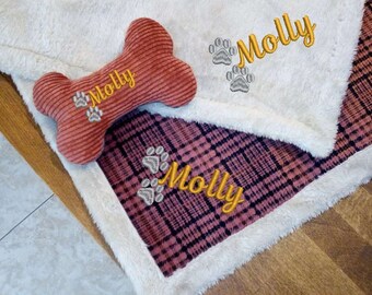 Könnte beinhalten: Ein Knochenspielzeug und eine Decke mit dem Namen "Molly" in Gold gestickt. Die Decke hat ein rosa-schwarzes Karomuster und einen weißen, flauschigen Rand. Das Spielzeug ist rotbraun.