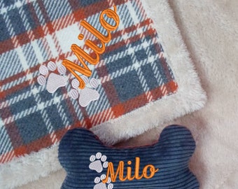 Könnte beinhalten: Ein Hunde-Set mit Decke und Spielzeug. Die Decke hat ein kariertes Muster in Grau, Orange und Weiß, mit dem Namen "Milo" in Orange gestickt, sowie Pfotenabdrücke. Das passende Spielzeug ist knochenförmig, marineblau und ebenfalls mit "Milo" und Pfotenabdrücken bestickt.
