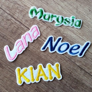 Könnte beinhalten: Vier gestickte Namensaufnäher: "Marysia" in Grün, "Lana" in Rosa, "Noel" in Blau und "Kian" in Gelb. Jeder Name ist auf einem weißen Filzgrund mit kontrastierendem Rand gestickt. Die Aufnäher werden auf einer Holzoberfläche präsentiert.