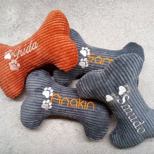 May include: Four corduroy dog bone toys in shades of orange and gray. Each bone is embroidered with a pet's name and paw prints. The names are "Frida", "Sam", "Anakin", and "Smudo".