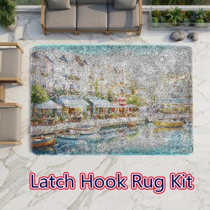 May include: A rectangular latch hook rug kit featuring a textured, pointillist-style scene of a European waterfront village with buildings, outdoor cafe seating, and small boats floating on the water. The text Latch Hook Rug Kit is printed below.