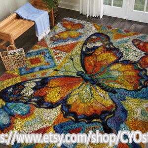 Op de afbeelding: Een kleurrijk tapijt met een vlindermotief in oranje, geel en blauw. De achtergrond heeft een geometrisch patroon. De Etsy shop URL is zichtbaar aan de onderkant.