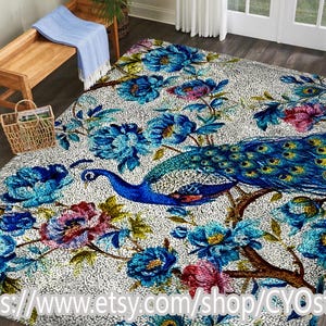 Puede incluir: Una alfombra grande con un diseño de pavo real. La alfombra presenta un pavo real con cuerpo azul y plumas de cola, rodeado de patrones florales azules y rosas sobre un fondo blanco. La alfombra es rectangular.