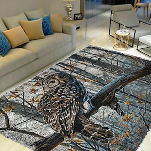 Op de afbeelding: Een grijs en blauw vloerkleed met een bruine uil die op een tak zit. De uil heeft bruine en witte veren en gele ogen. Het vloerkleed heeft een bosontwerp met takken en bladeren.