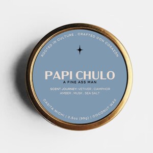 May include: A round gold-toned metal candle tin with a blue label. The text reads PAPI CHULO, A FINE ASS MAN, ROOTED IN CULTURE, CRAFTED CON CORAZON, SCENT JOURNEY: VETIVER, CAMPHOR, AMBER, MUSK, SEA SALT, CASITA MICHI, 3.5oz (99g), COCONUT WAX.