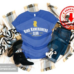May include: A royal blue t-shirt with a gold graphic design that says "King Kamehameha Club Est. 1905" with a small gold silhouette of a person's head with a crown on top. The t-shirt is laid out on a blanket with a pair of black ankle boots and blue jeans.
