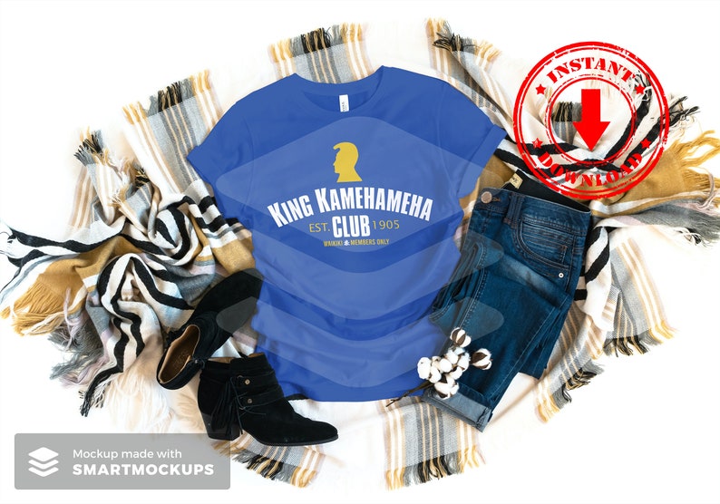 May include: A royal blue t-shirt with a gold graphic design that says "King Kamehameha Club Est. 1905" with a small gold silhouette of a person's head with a crown on top. The t-shirt is laid out on a blanket with a pair of black ankle boots and blue jeans.