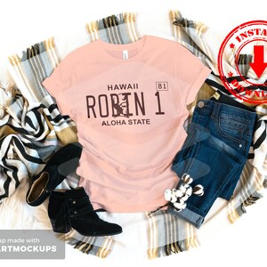 May include: A pink t-shirt with a brown graphic of a Hawaii license plate that says "Hawaii Robin 1 Aloha State". The shirt is laid out on a white background with a plaid blanket and blue jeans.