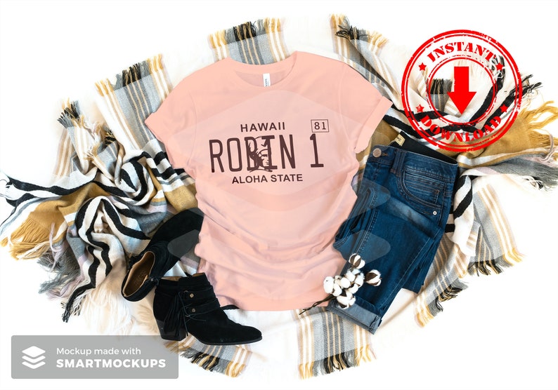 May include: A pink t-shirt with a brown graphic of a Hawaii license plate that says "Hawaii Robin 1 Aloha State". The shirt is laid out on a white background with a plaid blanket and blue jeans.
