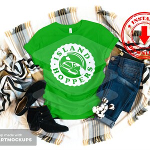 May include: A green t-shirt with a white graphic that says "Island Hoppers" with a cartoon car inside the circle. The shirt is laying on a blanket with a pair of black boots and blue jeans.