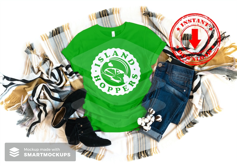 May include: A green t-shirt with a white graphic that says "Island Hoppers" with a cartoon car inside the circle. The shirt is laying on a blanket with a pair of black boots and blue jeans.