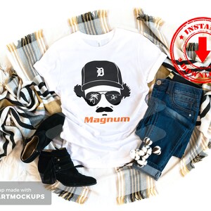 May include: White t-shirt with a black and white illustration of a person wearing a baseball cap with the letter "D" on it and sunglasses. The text "Magnum" is printed in orange below the illustration.