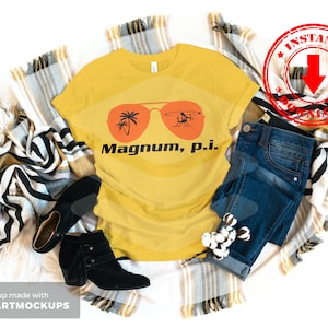 May include: A yellow t-shirt with a graphic of sunglasses and a helicopter with the text "Magnum, p.i." printed on it. The t-shirt is laid out on a blanket with a pair of black boots and blue jeans.