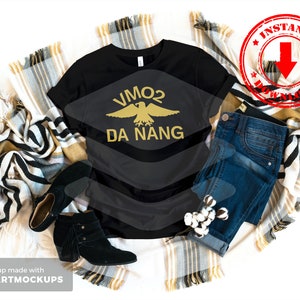 Puede incluir: Camiseta negra con un gráfico de águila dorada y el texto "VMO2 DA NANG". La camiseta se muestra sobre un fondo blanco con una manta y unos vaqueros.