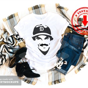 May include: White t-shirt with a black and white silhouette of a baseball player wearing a cap with the letter "D" on it. The t-shirt is laid out on a blanket with a pair of black boots and blue jeans.