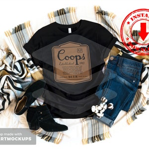 May include: A black t-shirt with a brown label that says "Coops Established 1917 Beer". The t-shirt is on a white background with a blanket and jeans.