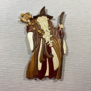 May include: A wooden carving of a wizard holding a staff and a ball of stars. The wizard is wearing a brown robe with a red sash and a pointed hat. The carving is painted in shades of brown, red, and gold.